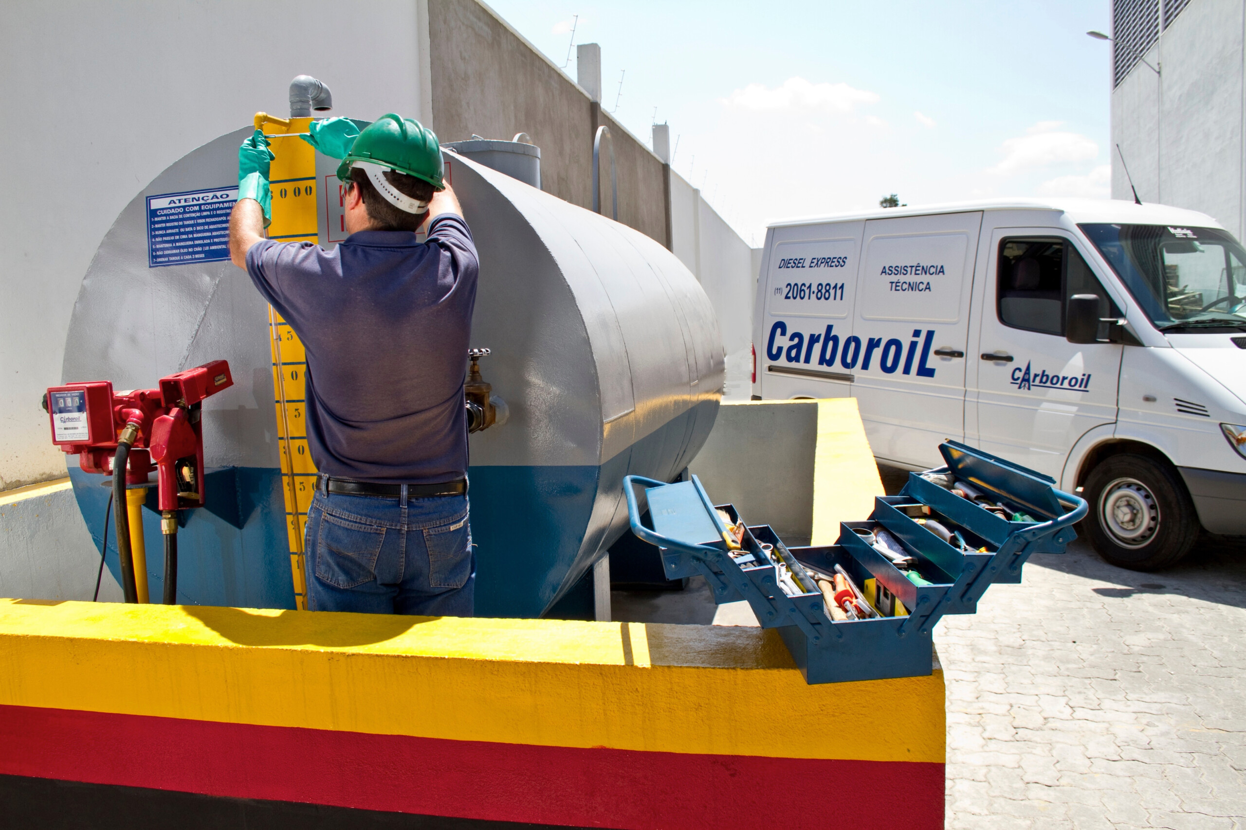 Profissional da Carboroil inspeciona tanque de diesel com equipamentos de medição, ao lado de van de assistência técnica, representando controle de qualidade do combustível.